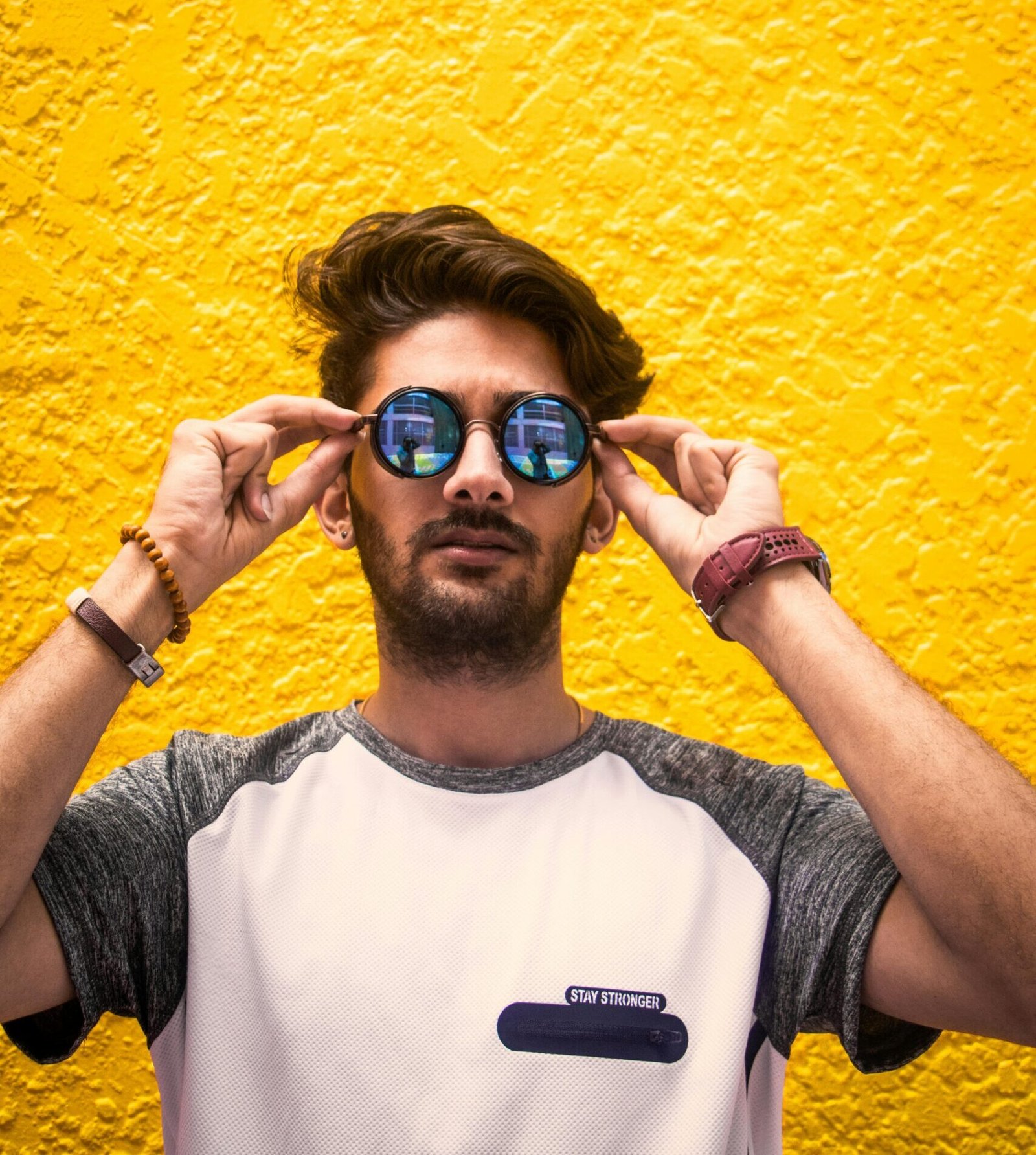 Young man with sunglasses standing against a vibrant yellow wall in daylight.